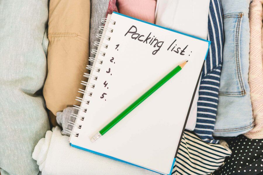 A notebook sitting on top of some clothes, with the words 'packing list' on it and a pencil