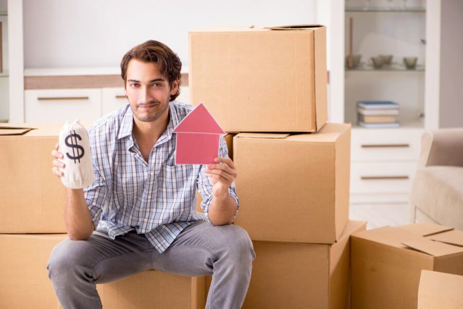 a man sitting in front of boxes, holding a dollar sign and a small house cut out, ready for moving cross-country