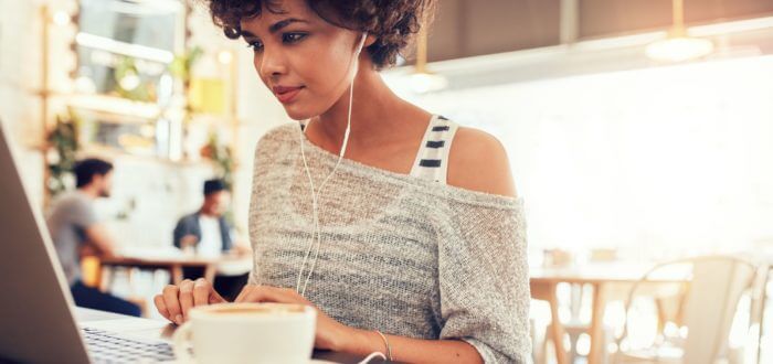 A girl with headphones, typing on the laptop in a cafe, a coffee on her left