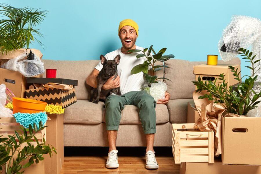 A man sitting on the couch, holding a dog and a plant, surrounded by plants and packages