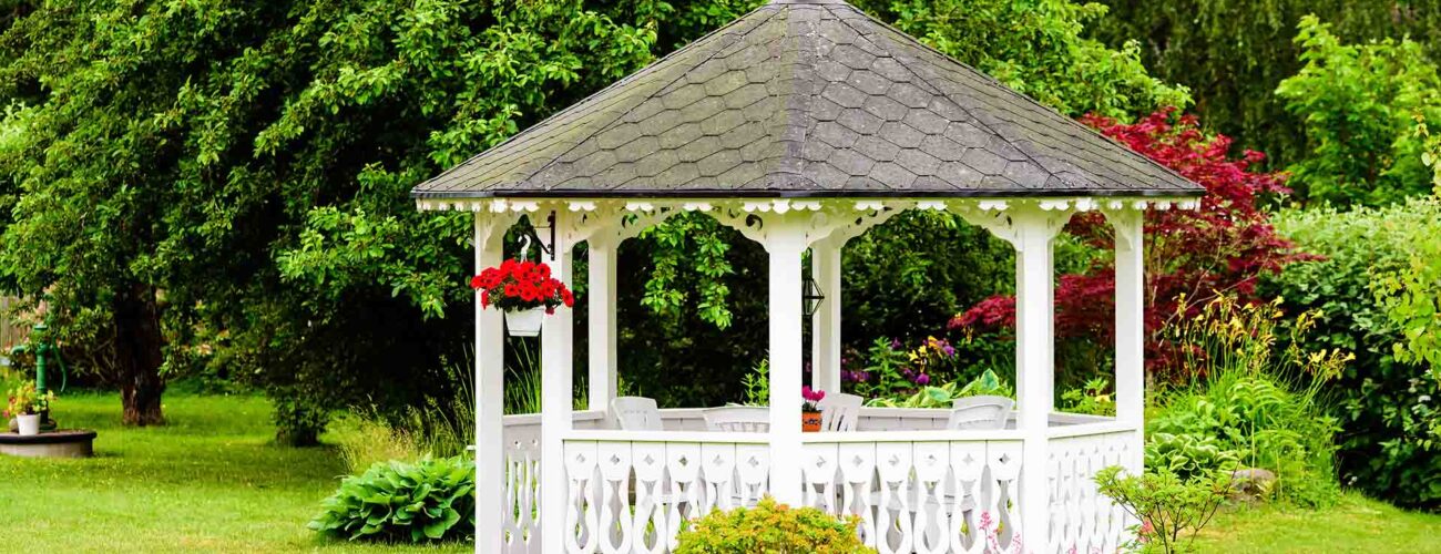 Lovely white gazebo with red flowers hanging from a basket. Trees and shrubs in the background. Fine garden with green grass.