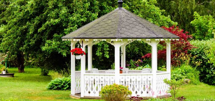 Lovely white gazebo with red flowers hanging from a basket. Trees and shrubs in the background. Fine garden with green grass.
