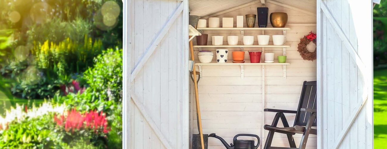 Garden shed filled with gardening tools. Shovels, rake, pots, water pitcher in storage hut. Green sunny garden in the background.